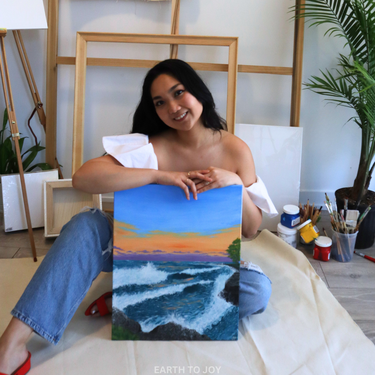 painter and artist smiling behind their art work and work of part. It is a painting of waves at lakeshore, artist is sitting with easels and canvases and paint tubs in the background