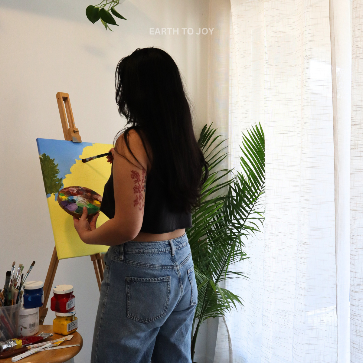 painter and artist brushing paint onto a canvas with an easel and paints in the background including greenery