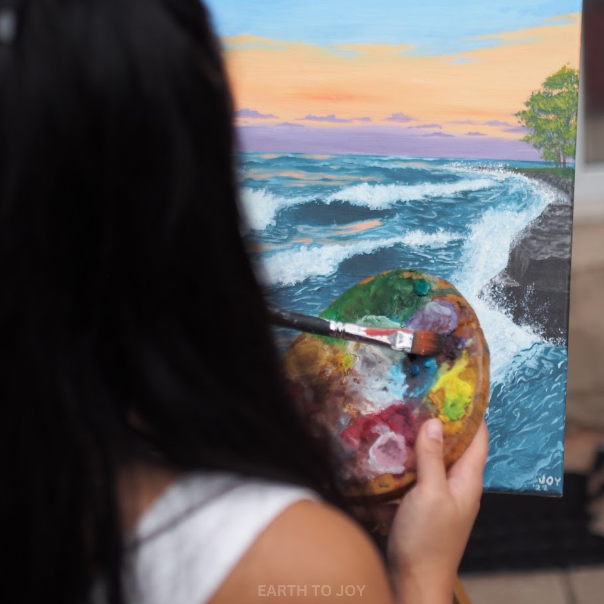 painter and artist brushing paint onto a paint palette with lots of pastel colours like purple, green, yellow, blue. Painting a lakeshore landscape