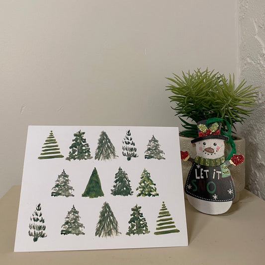 Multiple trees on a horizontal greeting card. The card is standing on a desk beside a snowman ornament infront of a succulent plant and a white wall