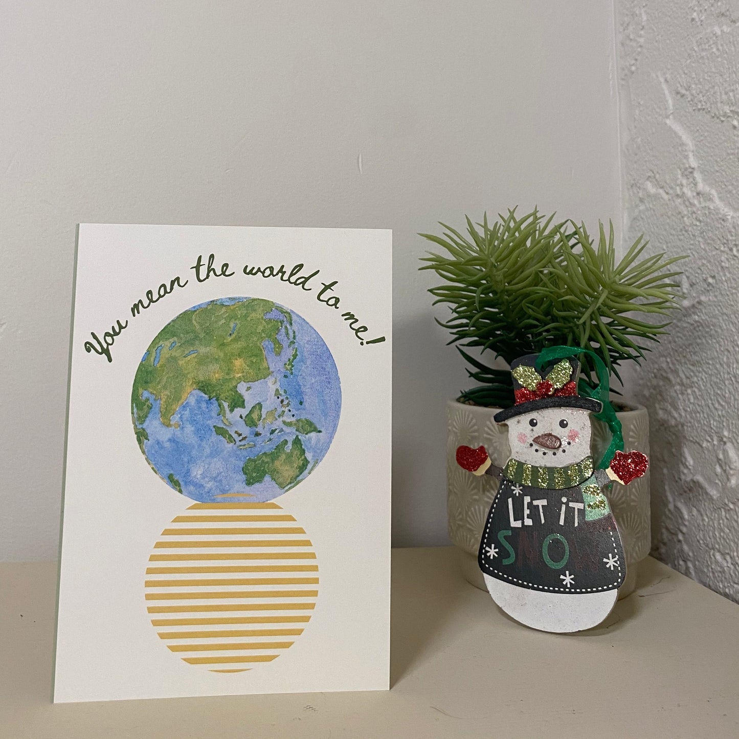 "You mean the world to me!" Affirmation card with a watercolour globe and a yellow striped circle below it. The card is standing on a desk beside a snowman ornament infront of a succulent plant and a white wall