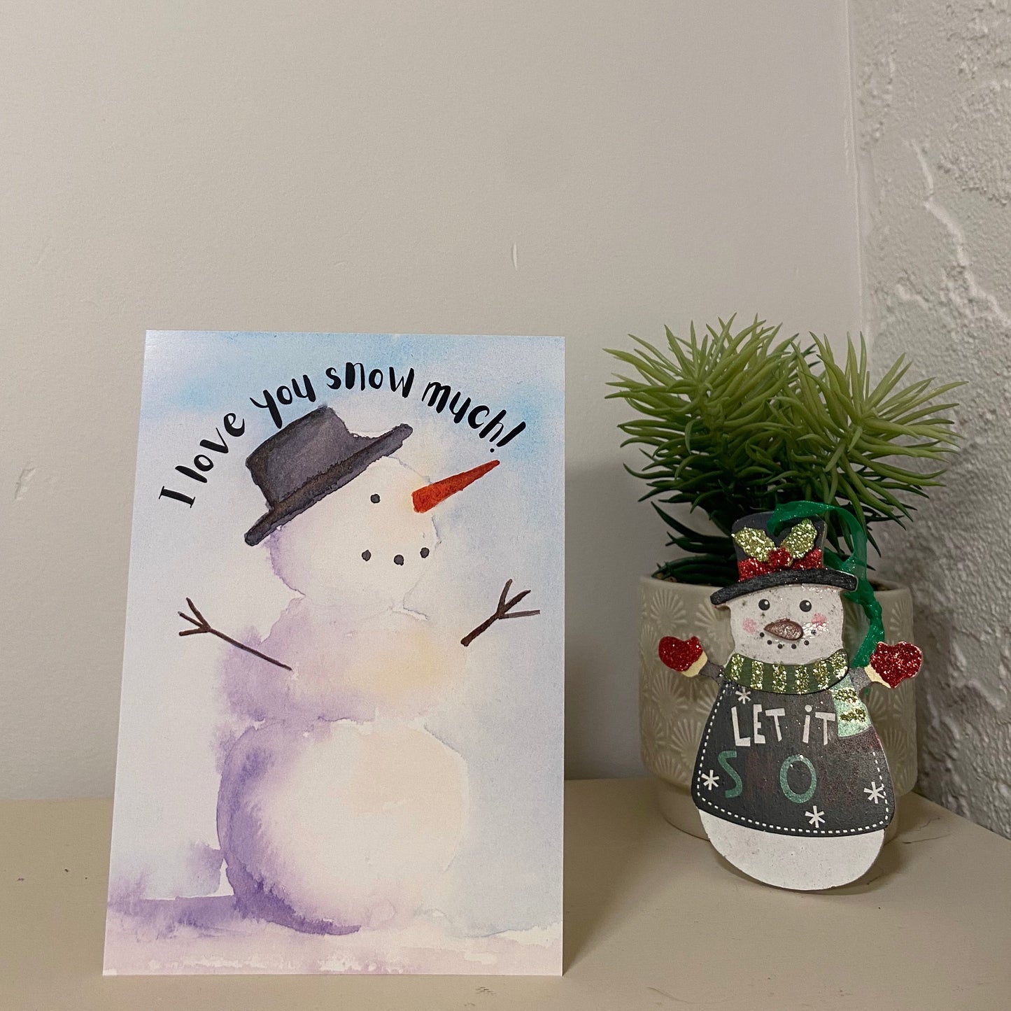 "I love you snow much!" holiday greeting card with watercolour winter snowman. The card is standing on a desk beside a snowman ornament infront of a succulent plant and a white wall