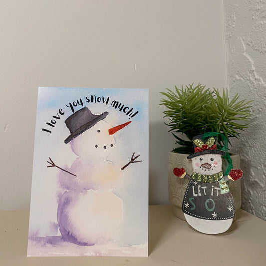 "I love you snow much!" holiday greeting card with watercolour winter snowman. The card is standing on a desk beside a snowman ornament infront of a succulent plant and a white wall